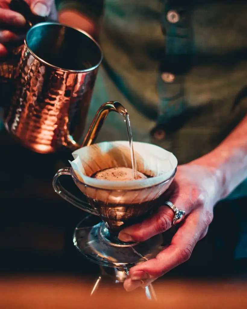 Two hands pouring from a hammered copper gooseneck kettle into a filter