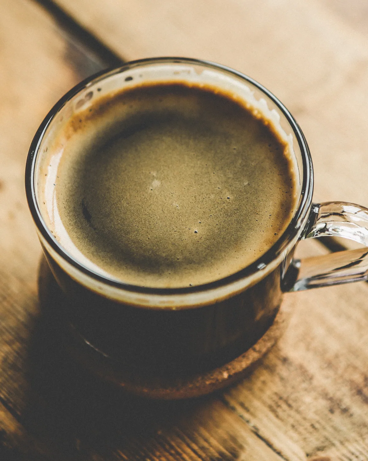 A glass mug of black coffee on a sunlit wooden table, seen from above