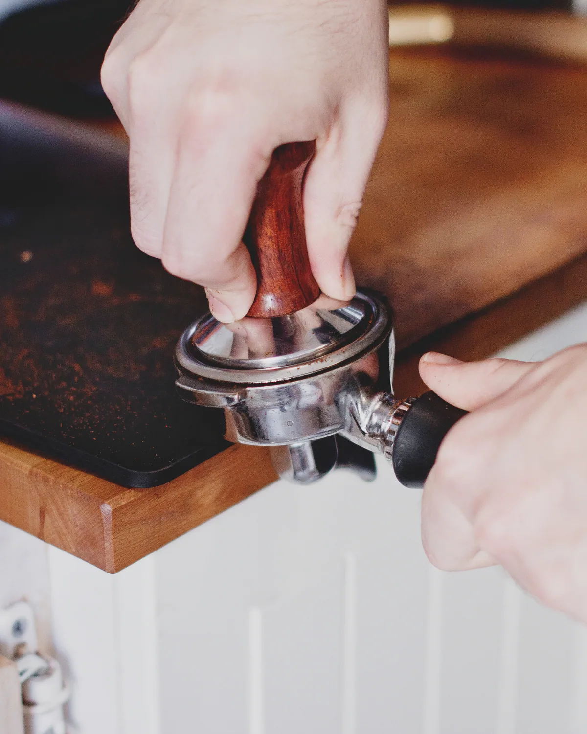 Two hands tamping ground coffee into a portafilter on a walnut station