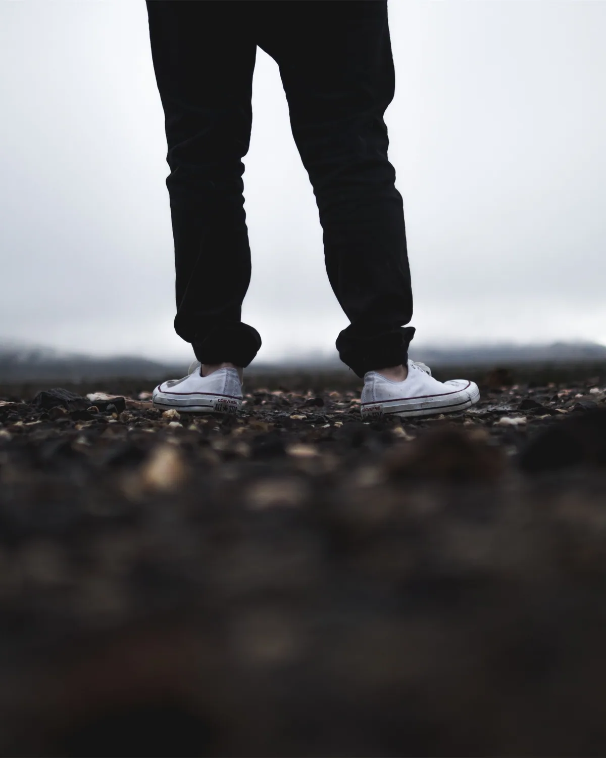 Legs in black jeans and white canvas sneakers standing on dark gravel