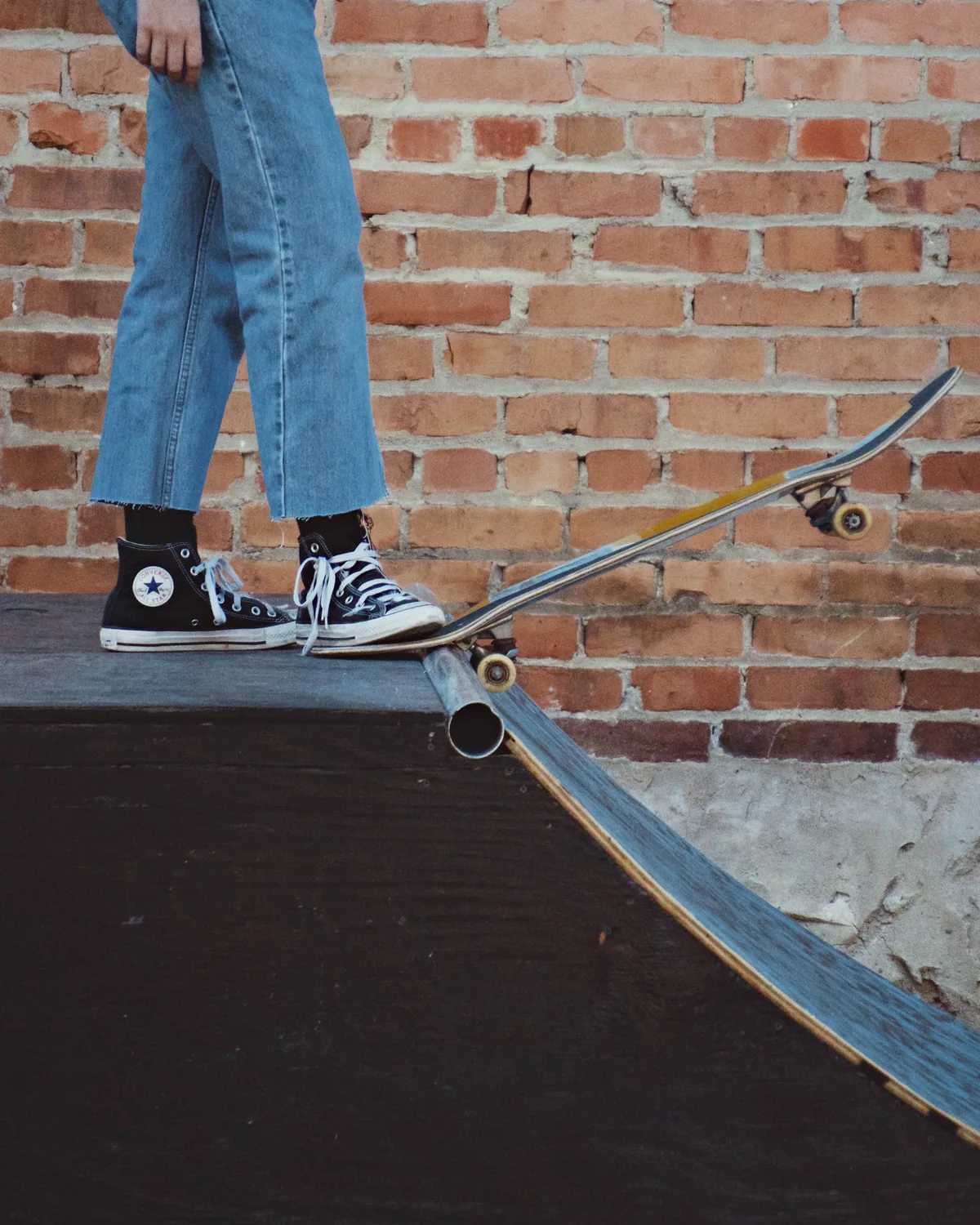 A skater in light-blue jeans standing on a plywood ramp against a brick wall