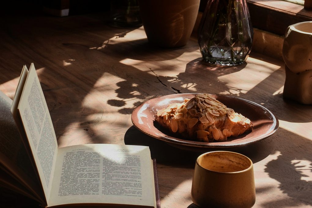 An open book, a croissant and a cup of coffee on a sunlit wooden table by the window, a slow start to the morning