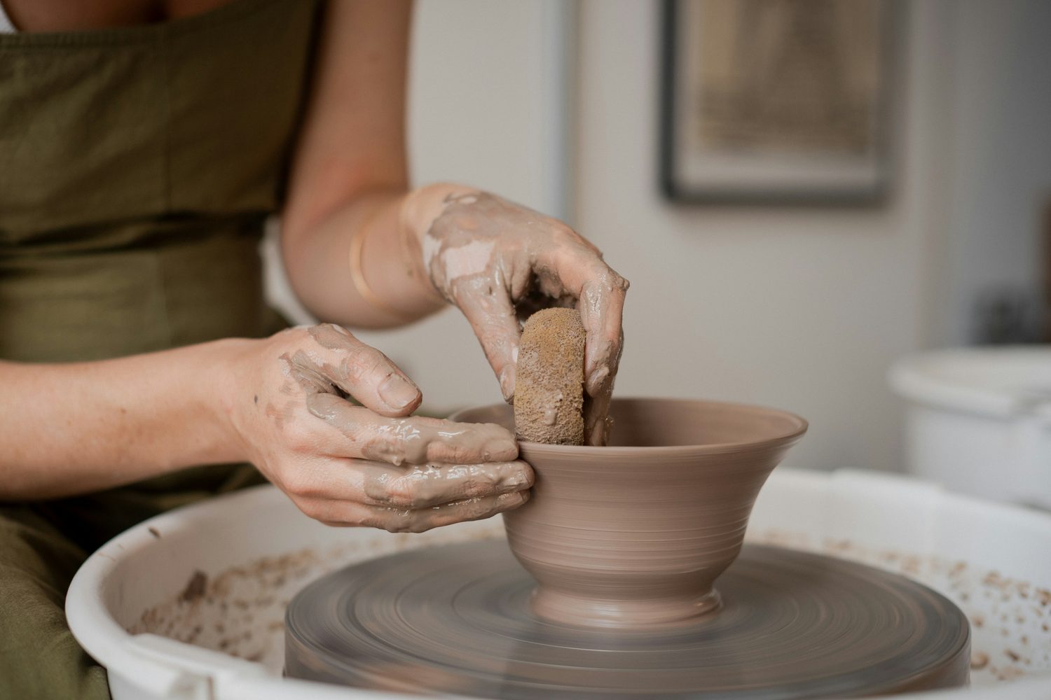 An adult learner in a work apron smoothing a bowl on the wheel with a sponge in a Bright Lane ceramics class
