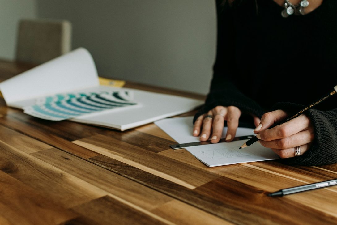 Mara Lindqvist sketching with a ruler at her walnut desk, a colour guide open beside her