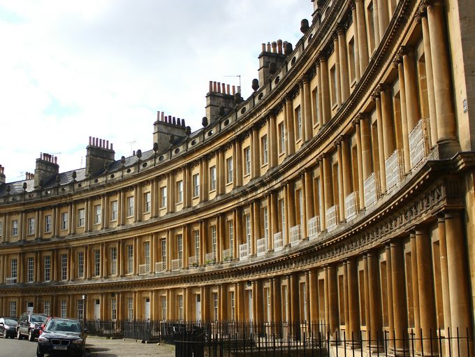 The curved Georgian facade of a Grade I listed Bath stone crescent with iron railings along the pavement