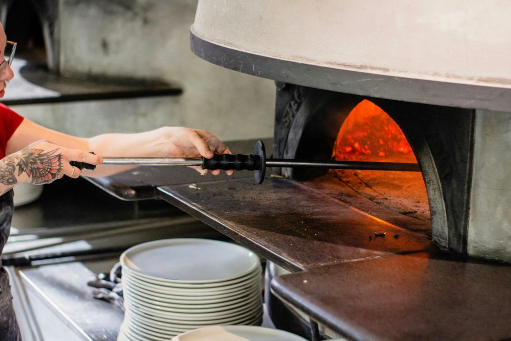 A cook working the wood-fired oven with a long peel while plates wait in a stack for service