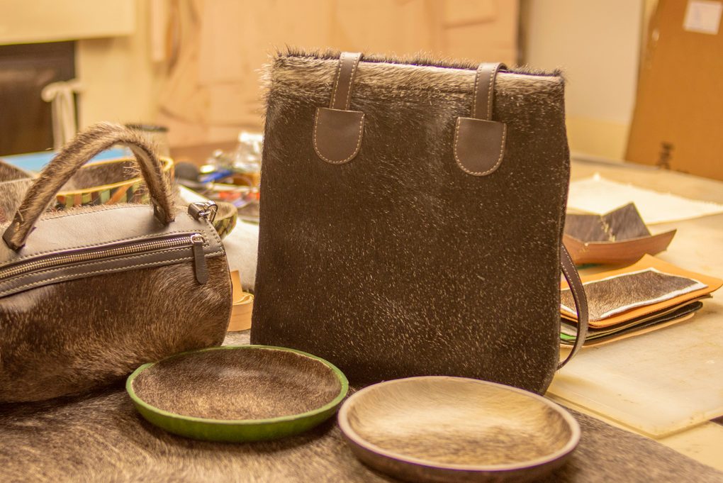 Two hair-on leather bags, a zip holdall and a tall tote, finished on the workbench beside a pair of leather trays in the Rowan & Reed workshop
