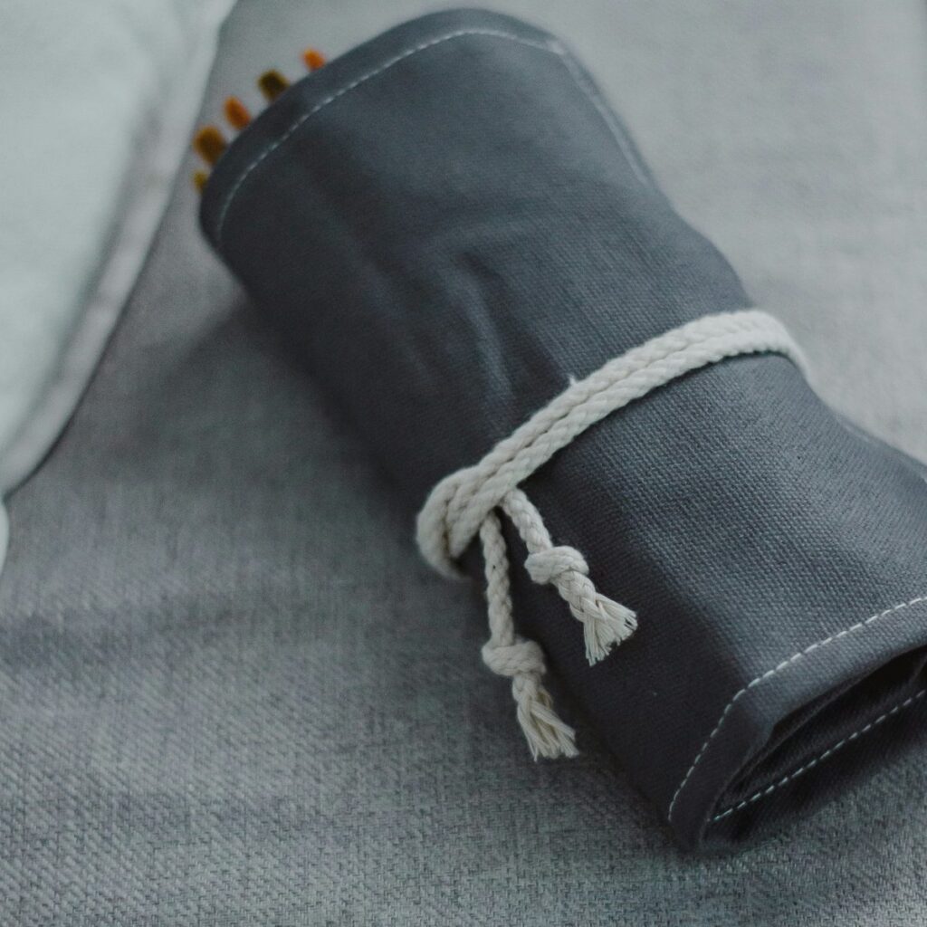 A slate grey canvas pencil roll tied with white cotton rope, resting on a grey linen cushion