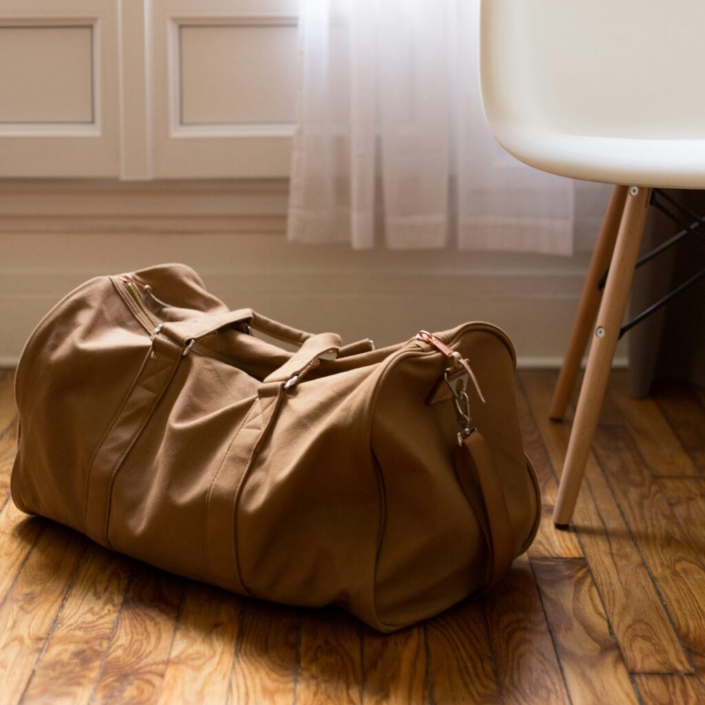 A tan canvas weekend holdall on a wooden floor beside a white chair in soft window light