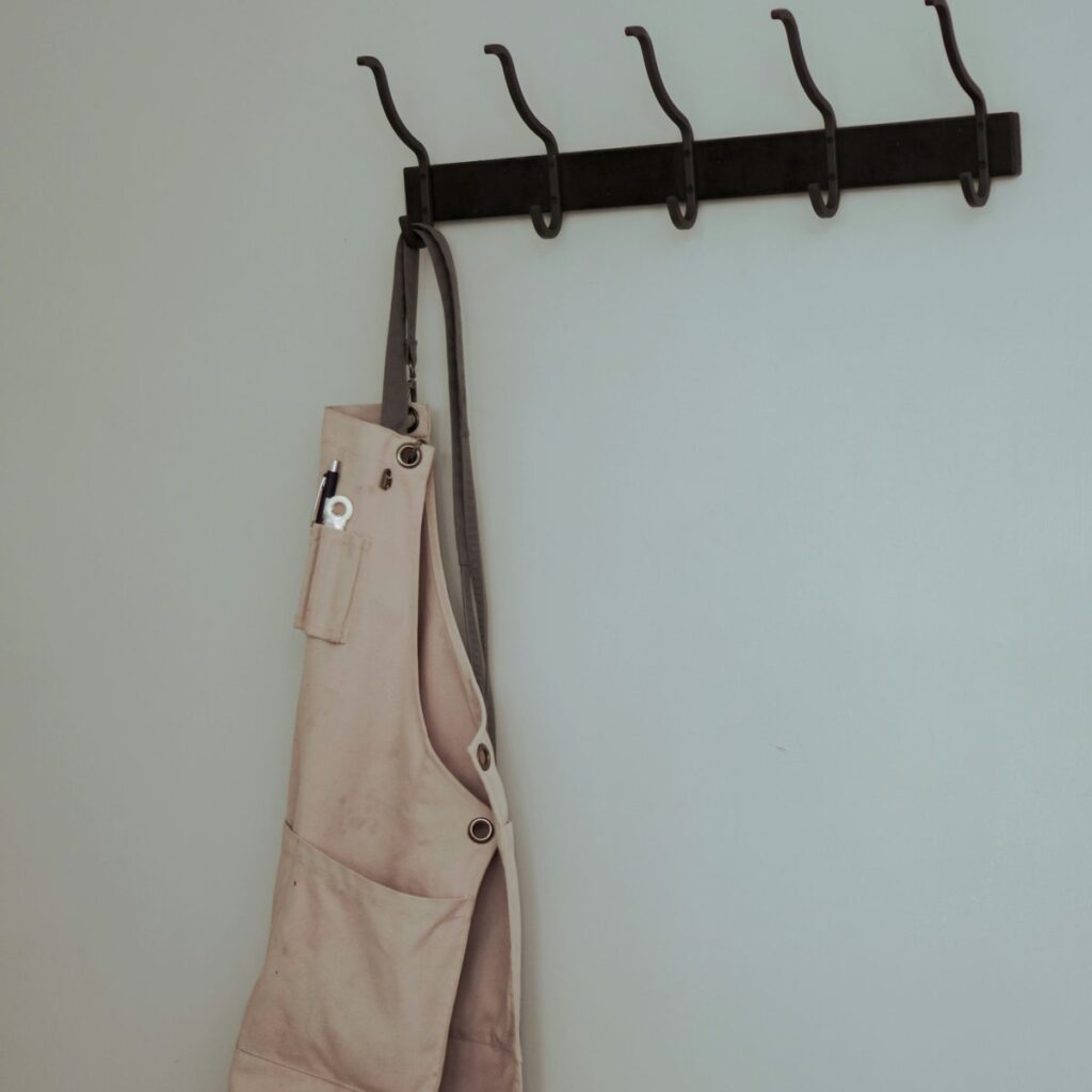 A pale canvas work apron with riveted pockets hanging from a black hook rail on a plain wall