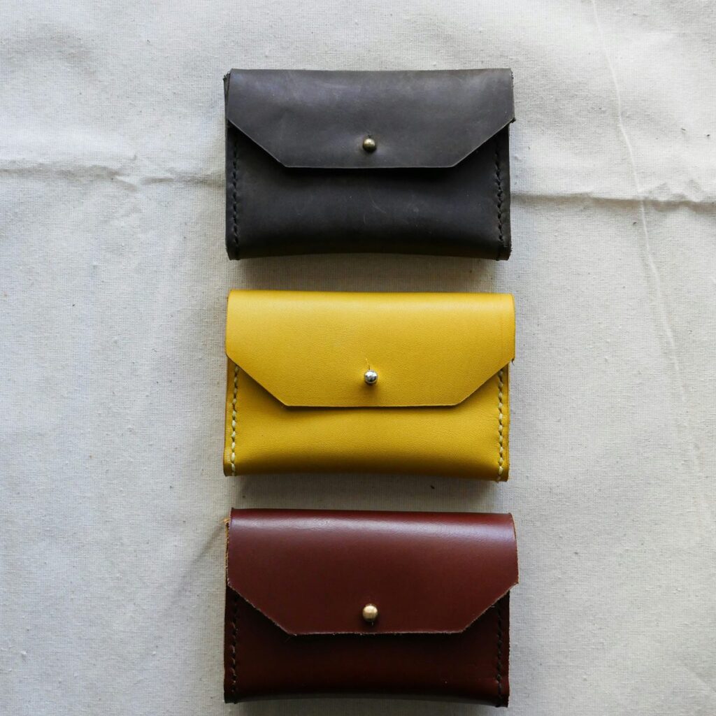 Three leather card pouches in slate, mustard and oxblood, each closed with a brass stud, laid on unbleached cotton