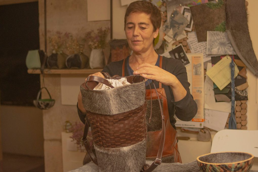 A maker in a leather apron fitting the drawstring lining into a woven leather bucket bag at the workbench, offcuts and pattern pieces pinned to the wall behind
