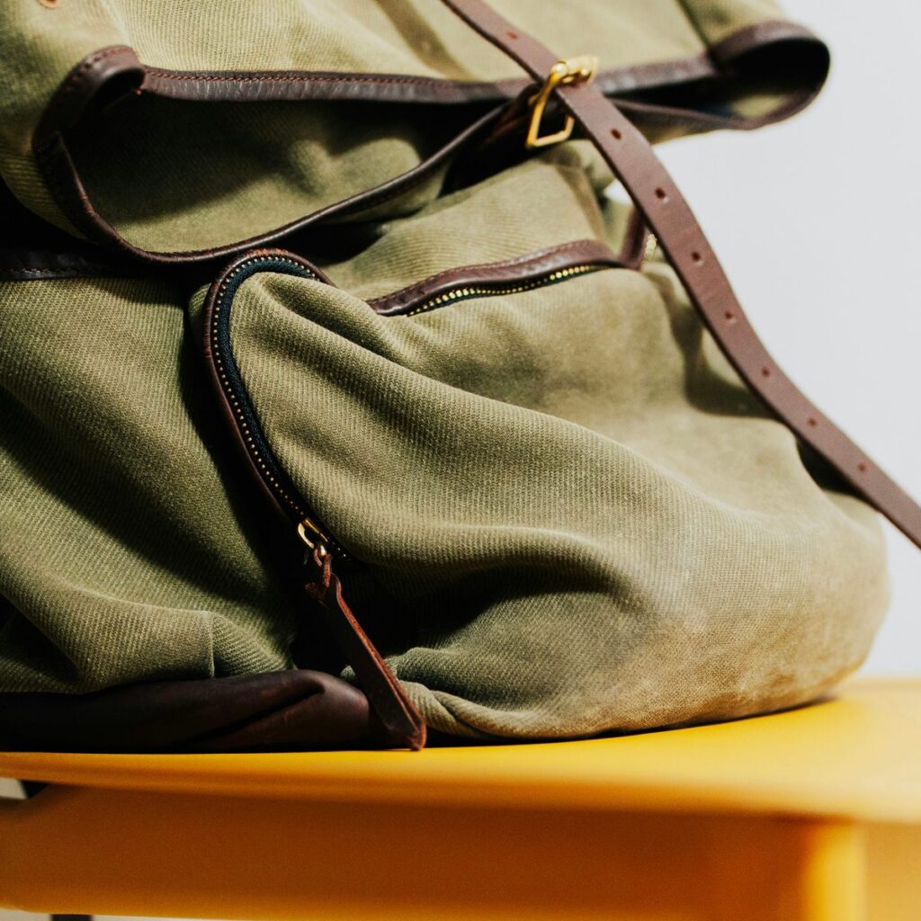 Close view of an olive waxed canvas rucksack with brown leather straps and a zipped front pocket resting on a chair