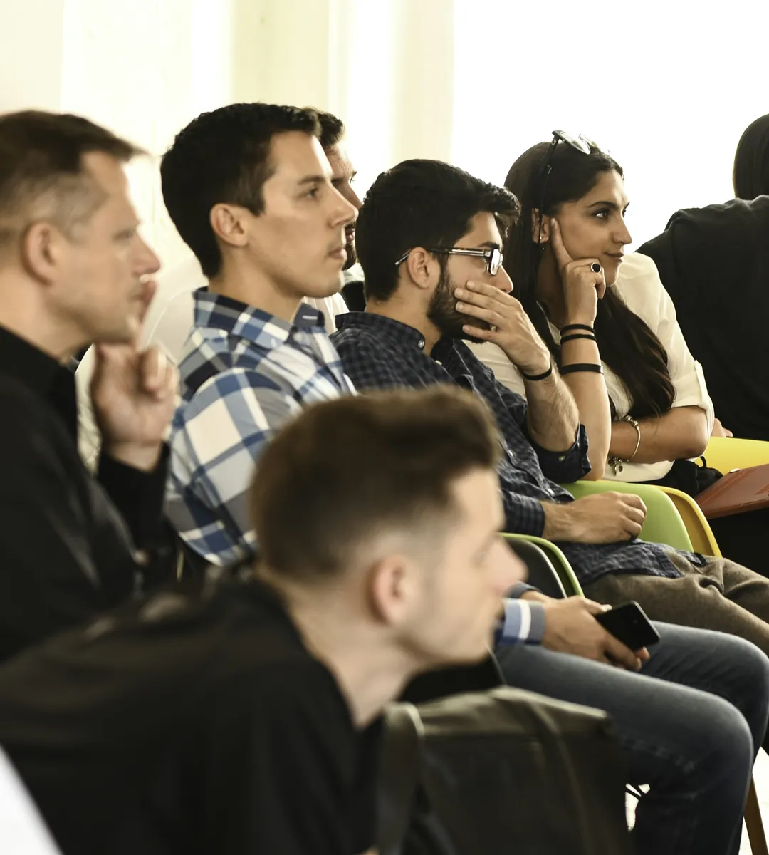 Attendees in a row of chairs listening to a talk: a man in a checked shirt looking ahead, a man in glasses with his hand to his chin, a woman resting her cheek on her hand