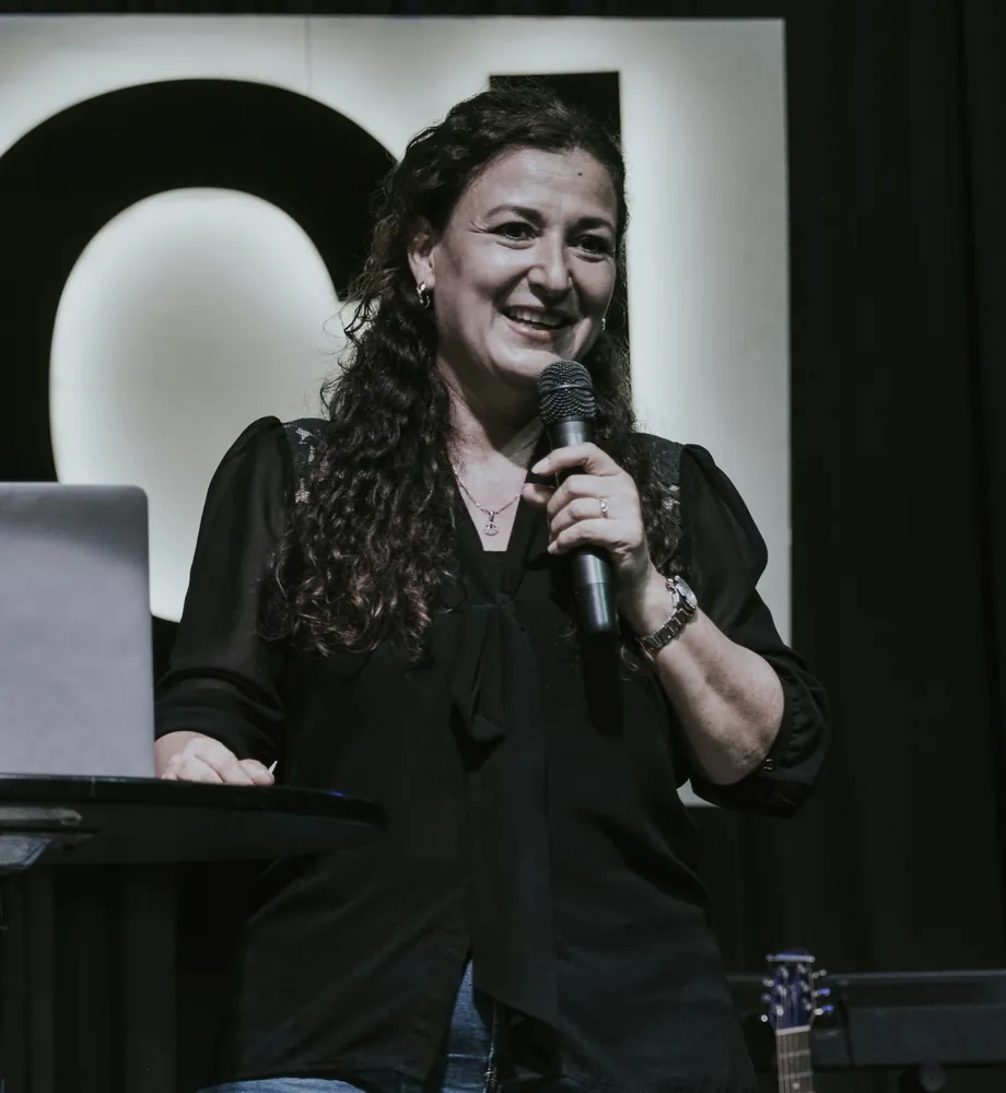Elena Ruiz laughing, holding a microphone, in a black blouse beside a lectern with a laptop