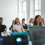 A workshop in a white room with tall windows, participants listening with notebooks behind a projector and an open laptop